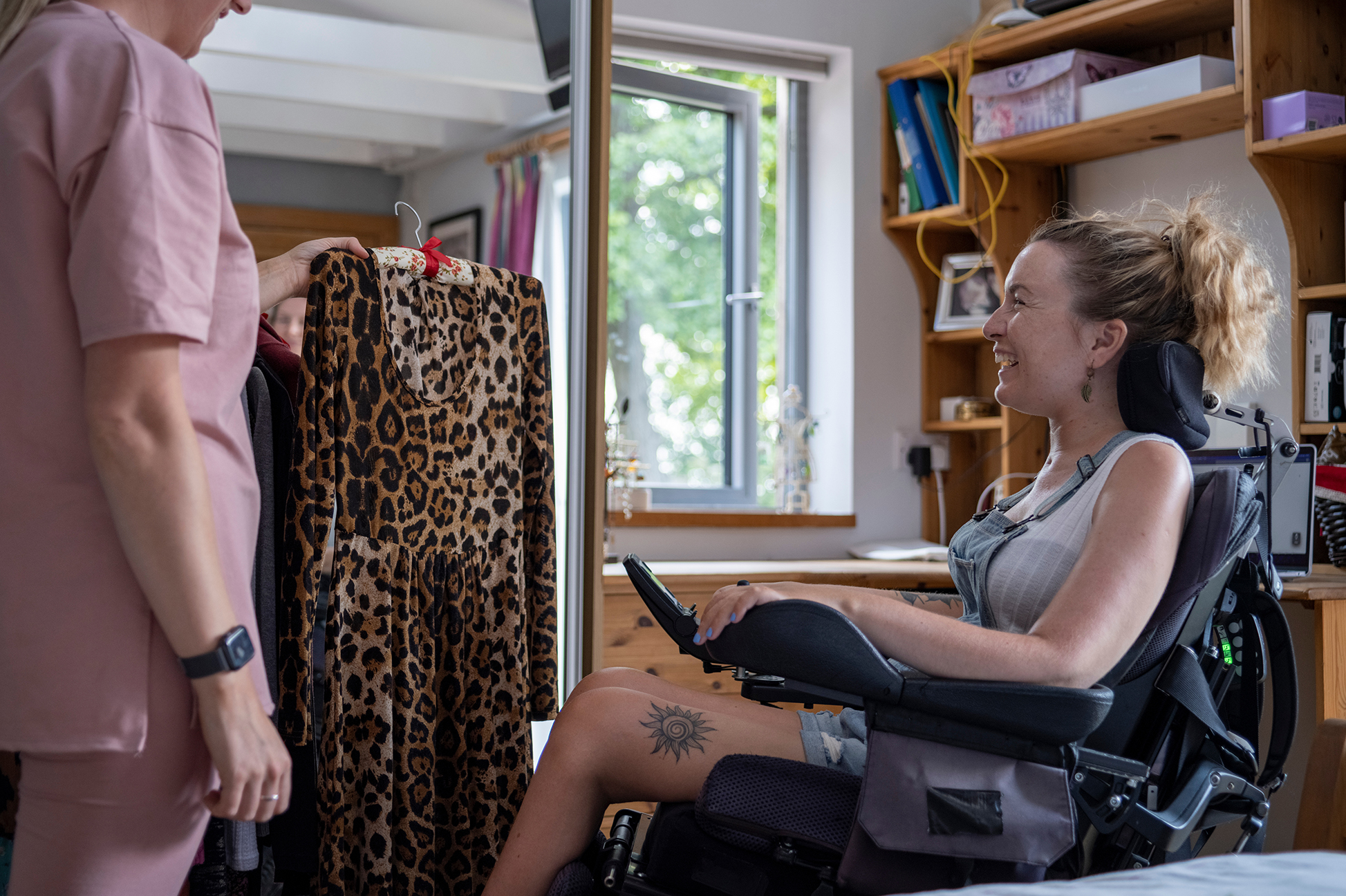 "A woman holds up an outfit to a woman sitting in a motorized wheelchair who is smiling"
