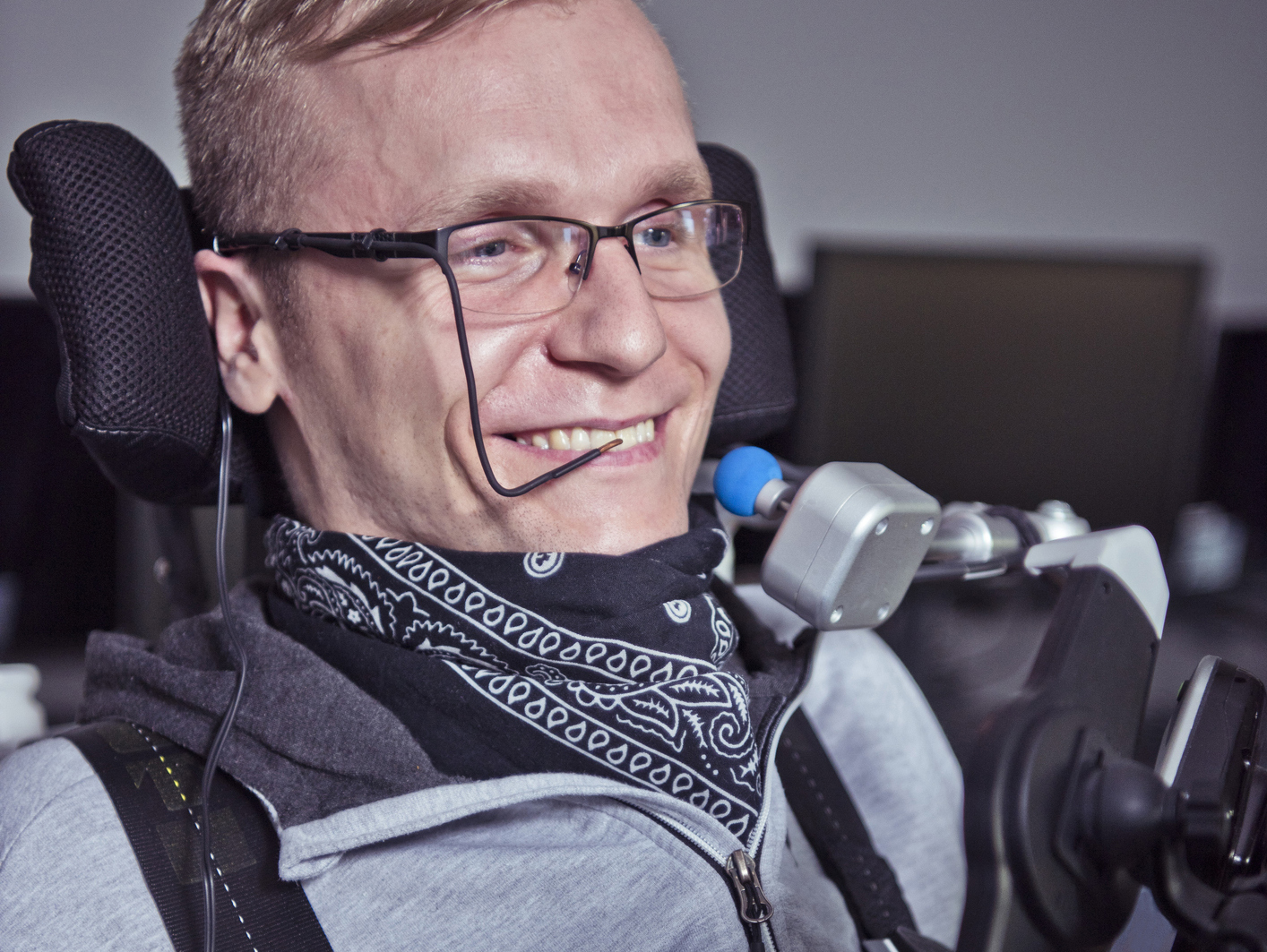 "A man with glasses seated in a wheelchair with assistive technology tools designed for accessibility smiles"