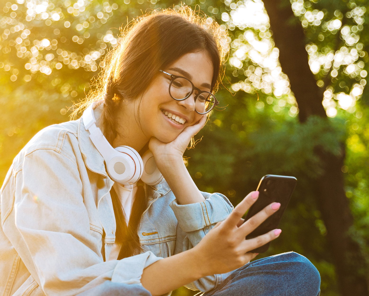 "A girl with glasses and headphones on her neck smiles as she looks at a smartphone while sitting outside"
