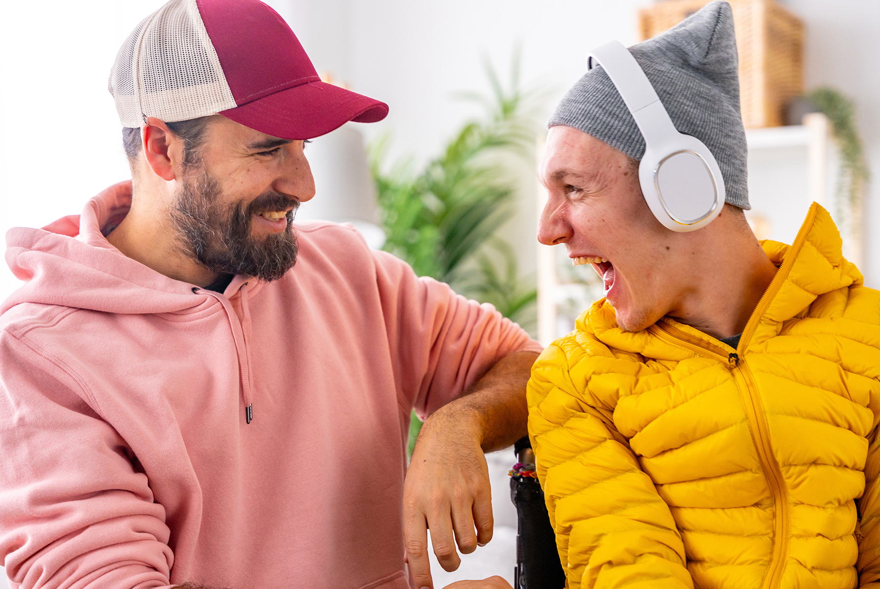 "A man wearing a ball cap and a pink hoodie smiles as he communicates with his friend who has ALS and is wearing a yellow jacket and headphones"