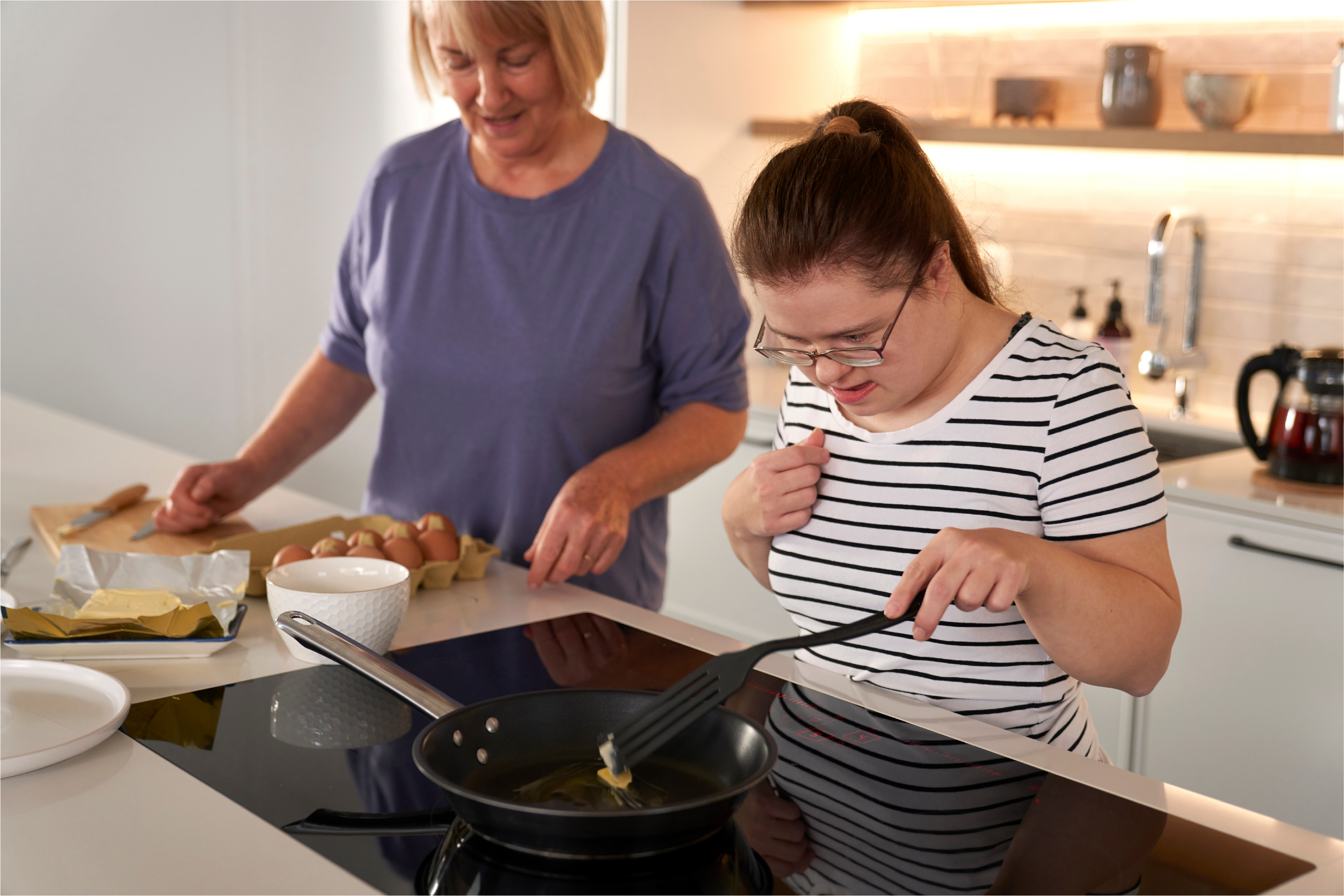 "An older woman with short hair helps a younger woman cooking at the stove"