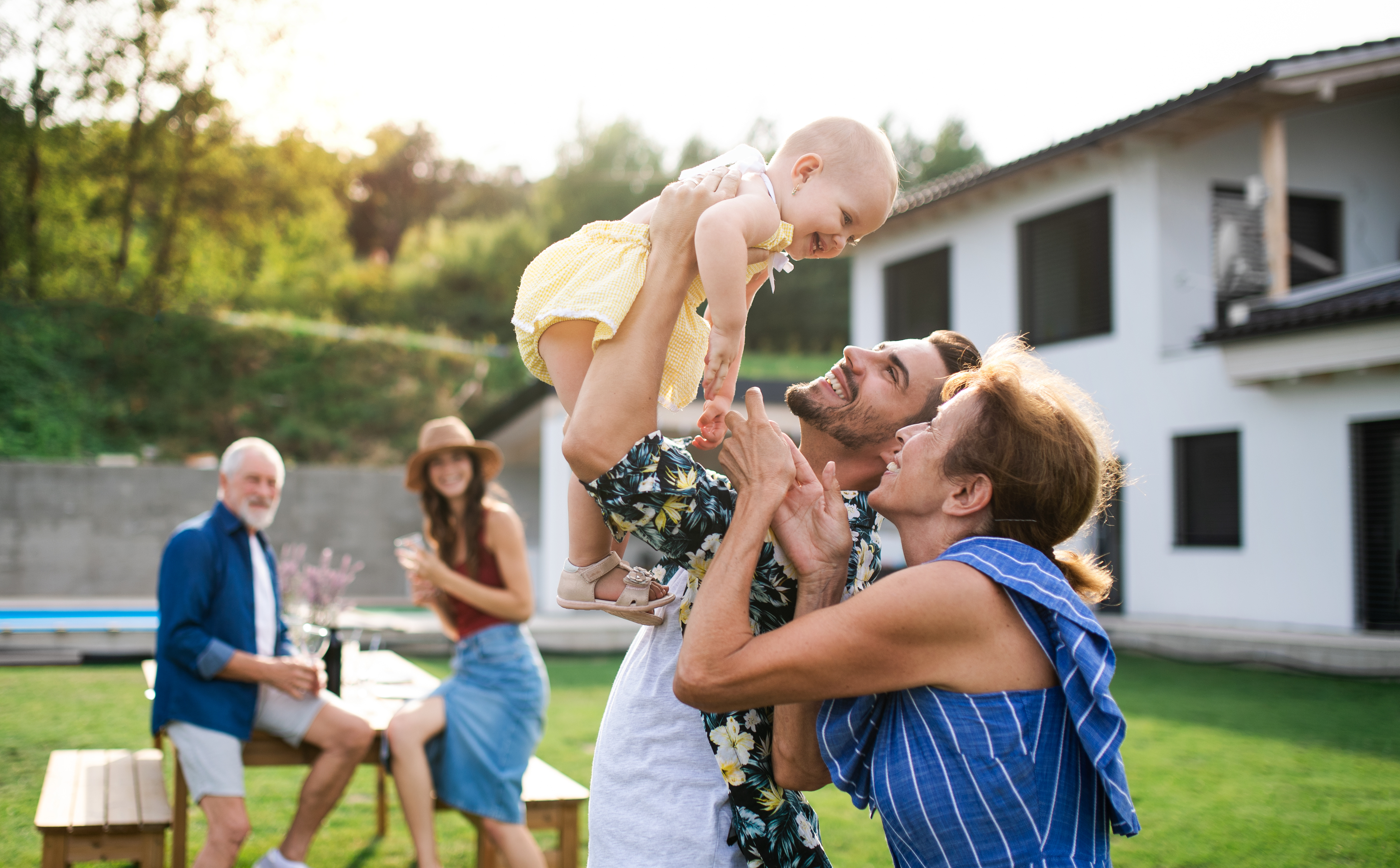 A family is in a backyard. Older family members are less clear in the background of the photo, while younger parents are holding up a young child