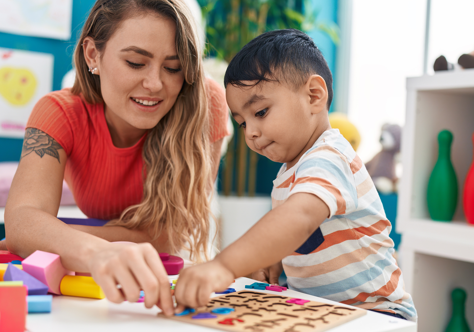 "A woman with long blond hair and a red shirt helps a young boy with dark hair and a striped shirt as they put together an alphabet puzzle at a table"