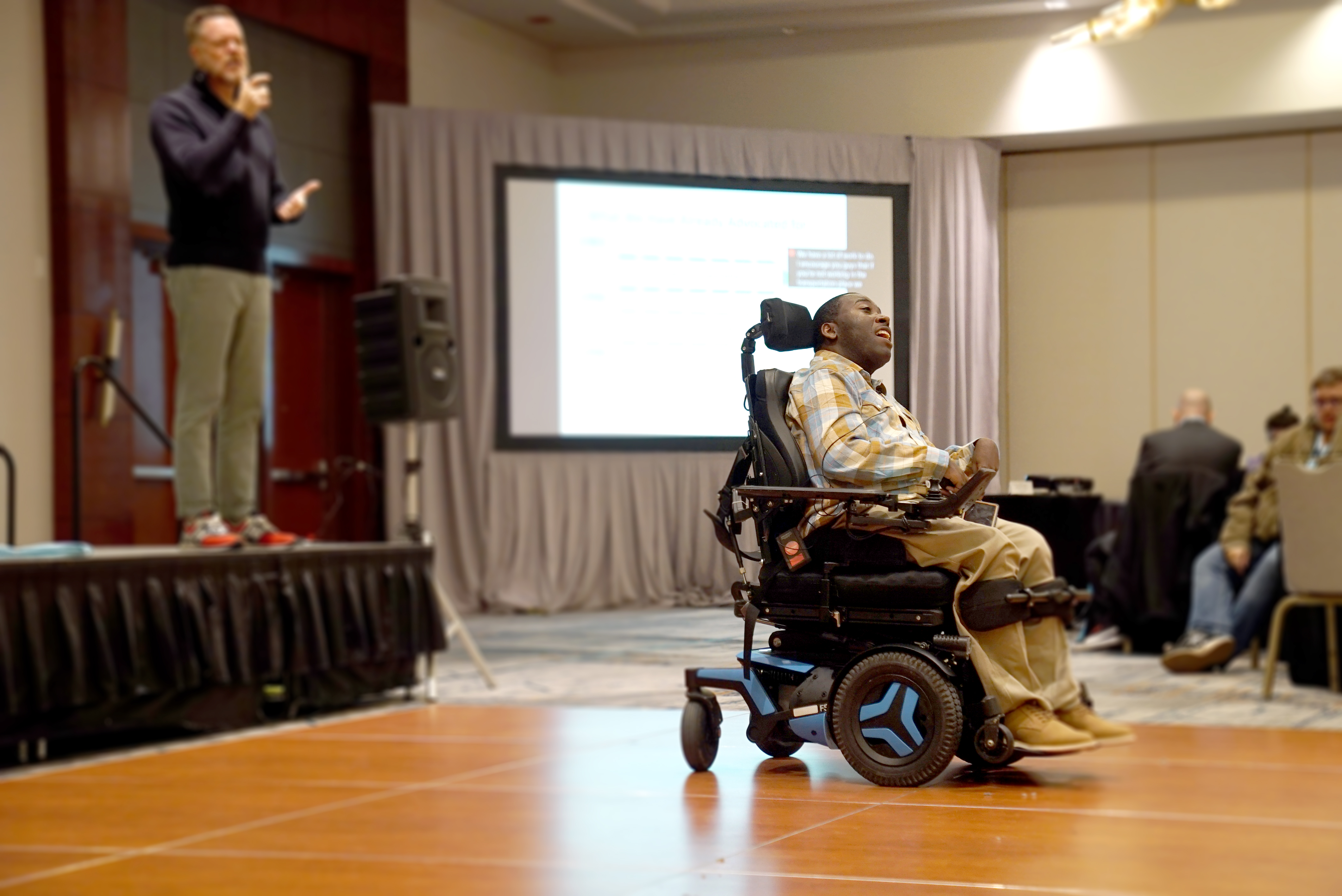 "A man with short black hair wearing a button up shirt and khaki pants while sitting in a motorized wheelchair speaks to an audience in a large conference room"