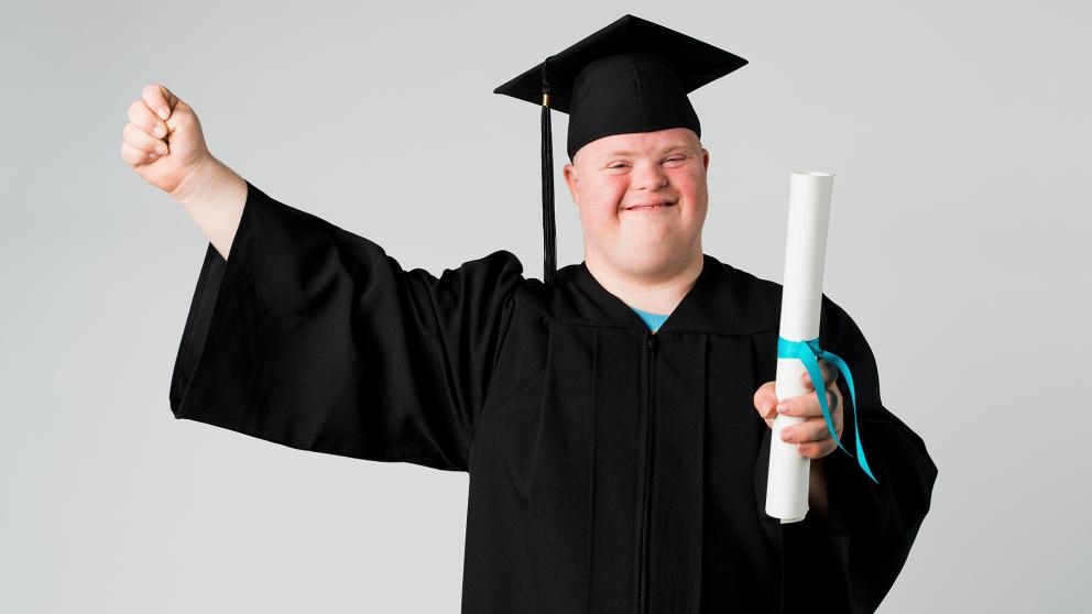 "A man with Down syndrome wearing a graduation outfit smiles and raises a fist in triumph"